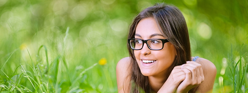 Young charming smiling dark-haired woman in glasses with black frame lies on green grass in summer city park.